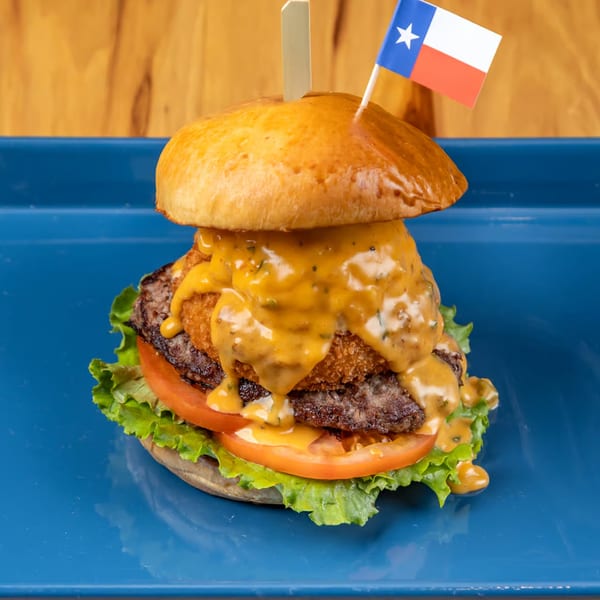 A LSA Burger Co. burger with a Texas flag pick, cheese sauce, onion rings, and lettuce.